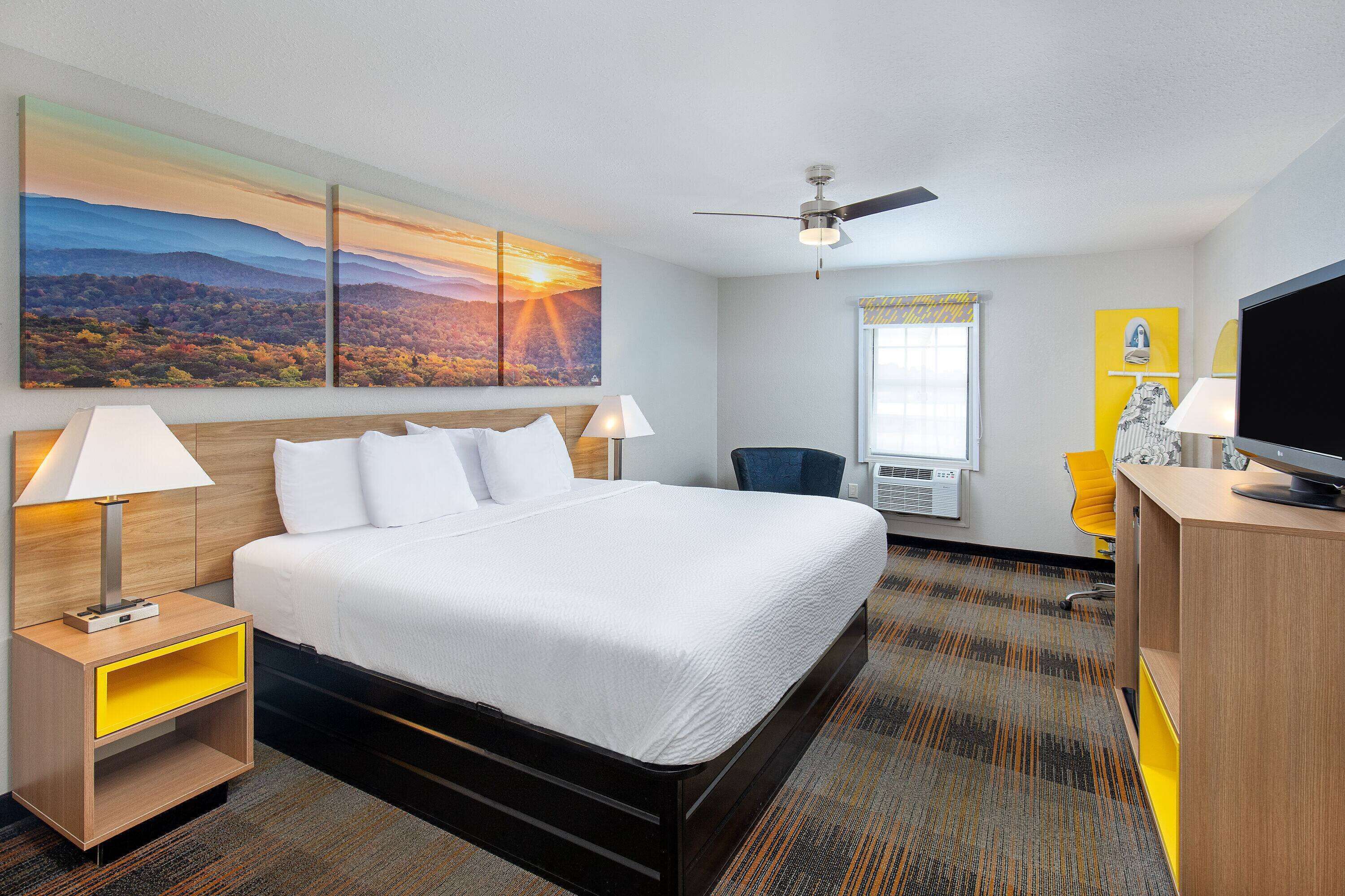 A hotel room with a bed, two bedside tables with lamps, a desk with a TV, a chair, and a large landscape painting on the wall.