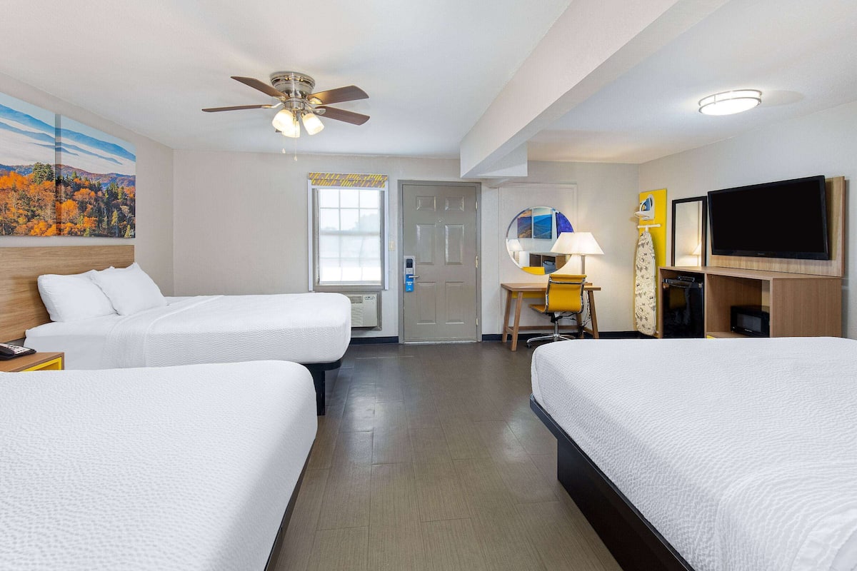 A hotel room with two beds, a ceiling fan, a TV, and a painting of autumn foliage.