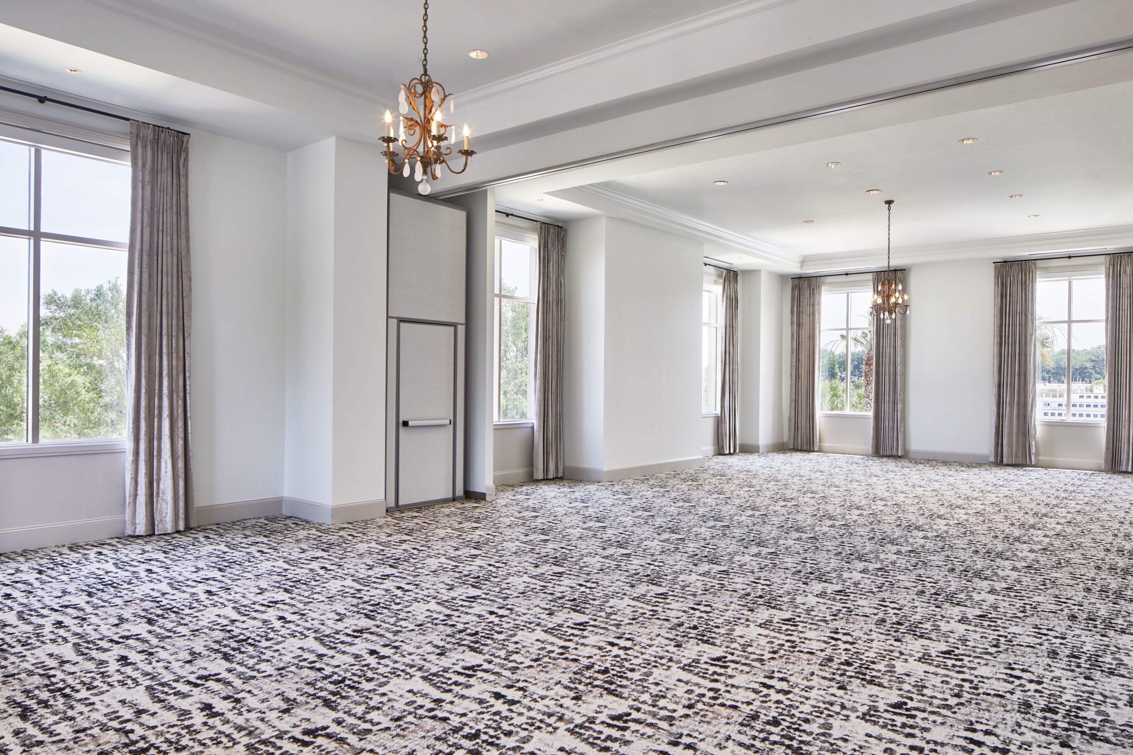 A spacious room with a patterned carpet, large windows with curtains, and a chandelier.