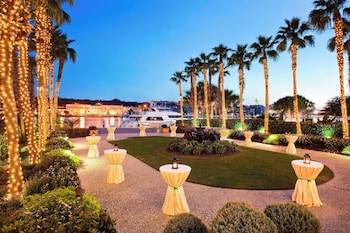 An aerial view of a resort with a central swimming pool surrounded by lounge chairs and umbrellas, palm trees, and a dock by the water. at The Westin Savannah Harbor Golf Resort & Spa