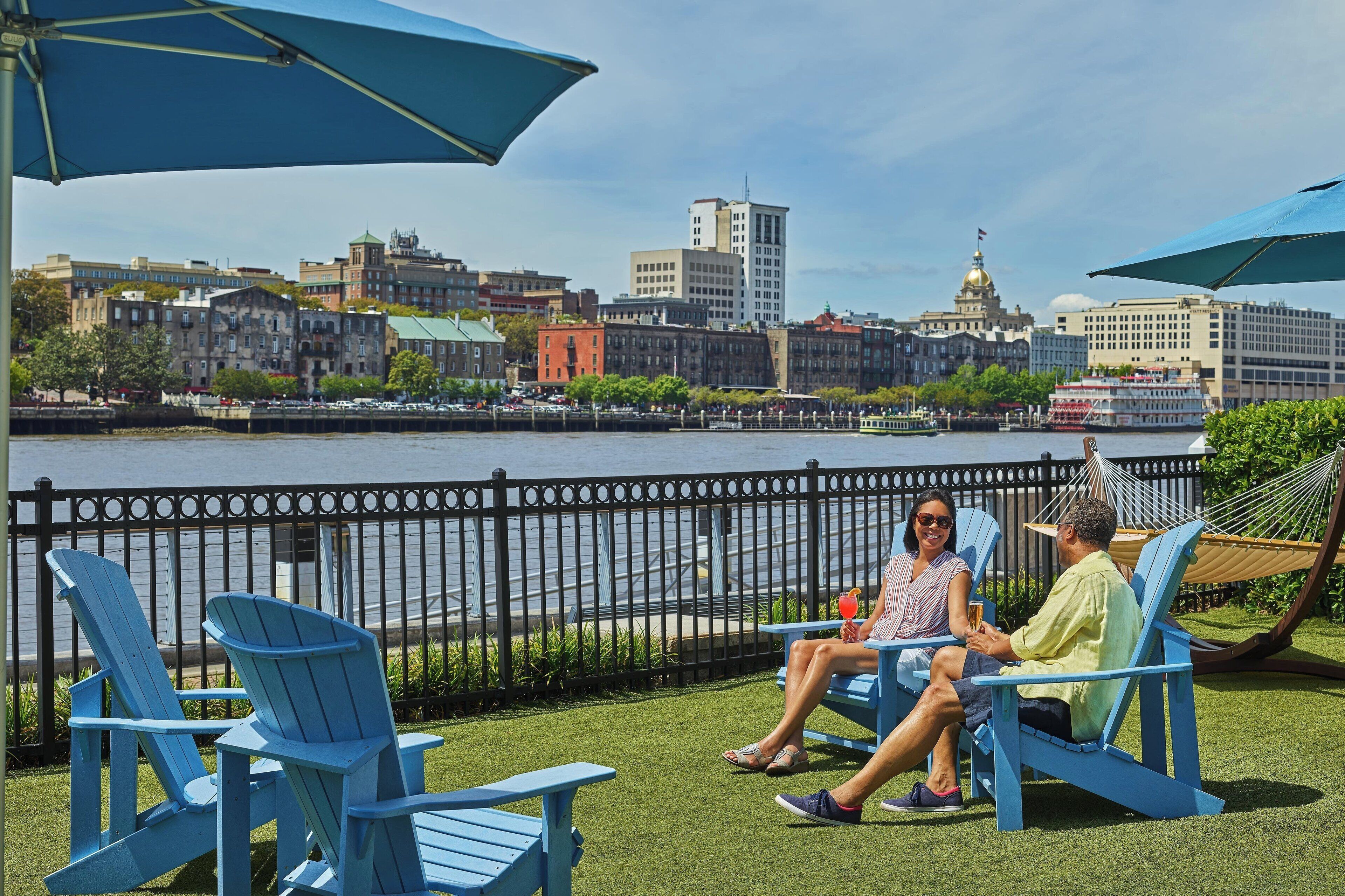 A waterfront patio with blue Adirondack chairs, a hammock, and a view of a city skyline across the river.
