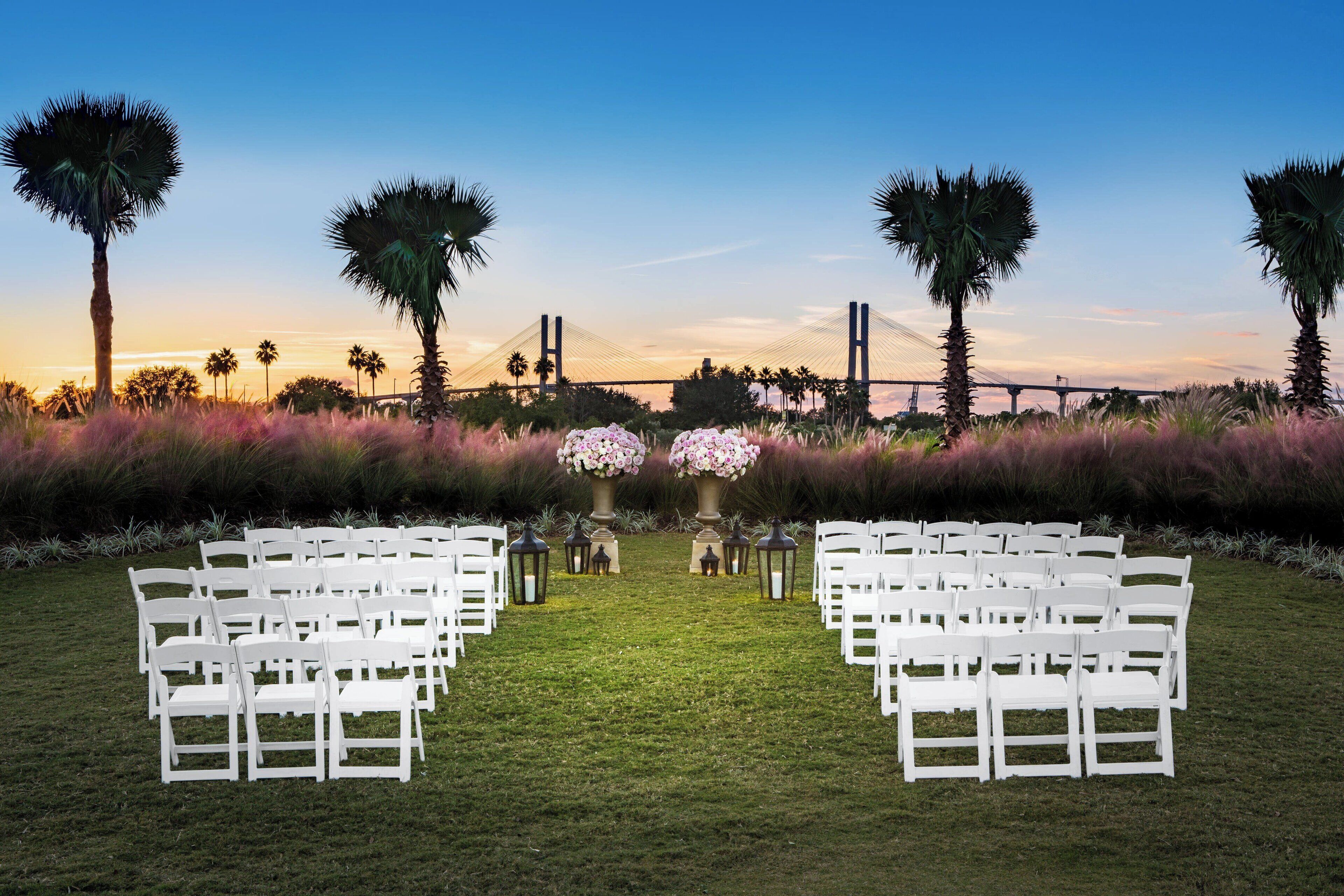 An outdoor wedding venue with white chairs arranged in rows, palm trees, and a bridge in the background.