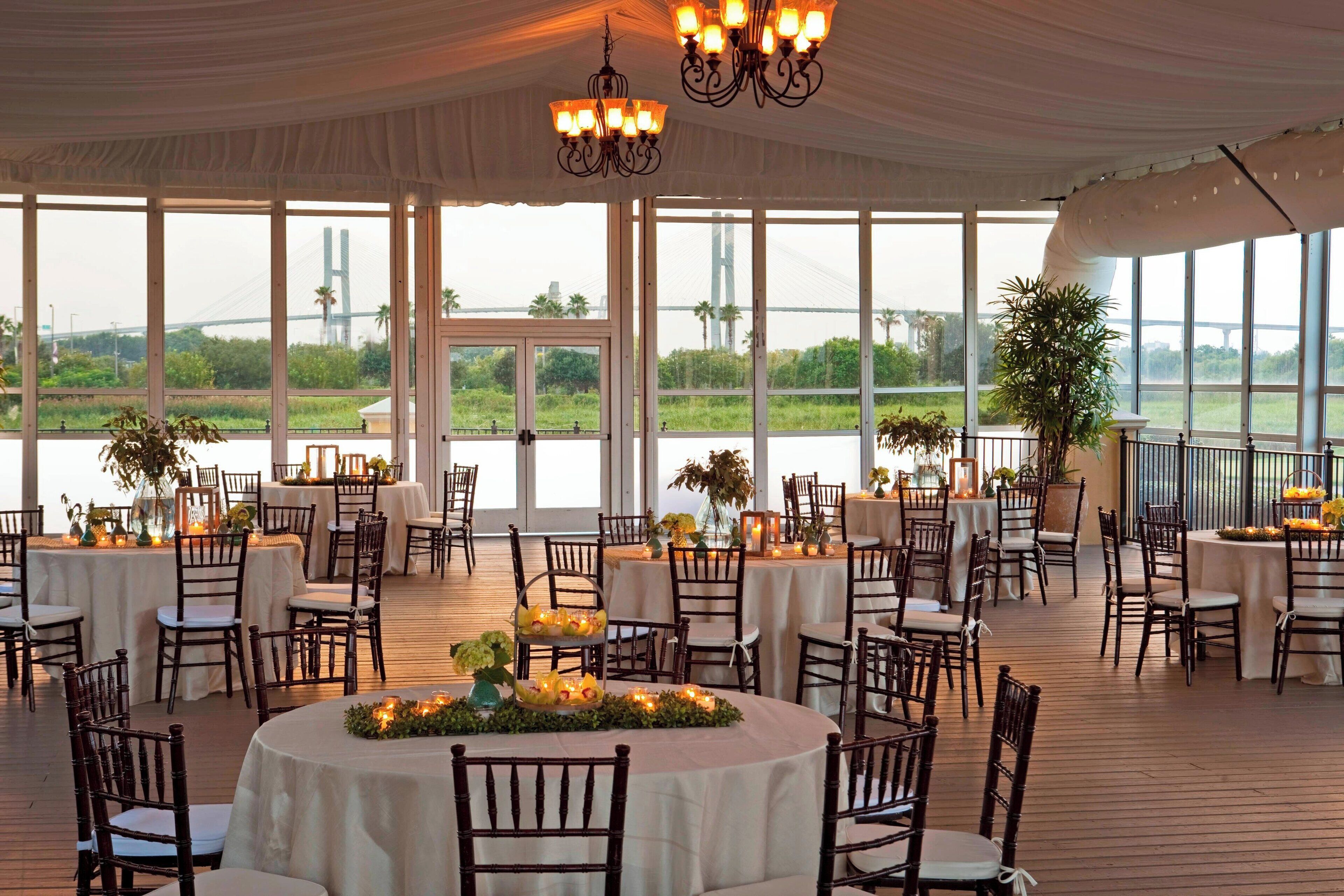 A spacious outdoor dining area with round tables and chairs, large windows offering views of a bridge and greenery.