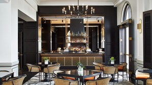 Eine Bar mit einer langen Theke, mehreren Flaschenregalen und einer Kristallleuchtre. Mehrere Korbstühle und ein runder Tisch mit einem Blumenstrauß stehen bereit.