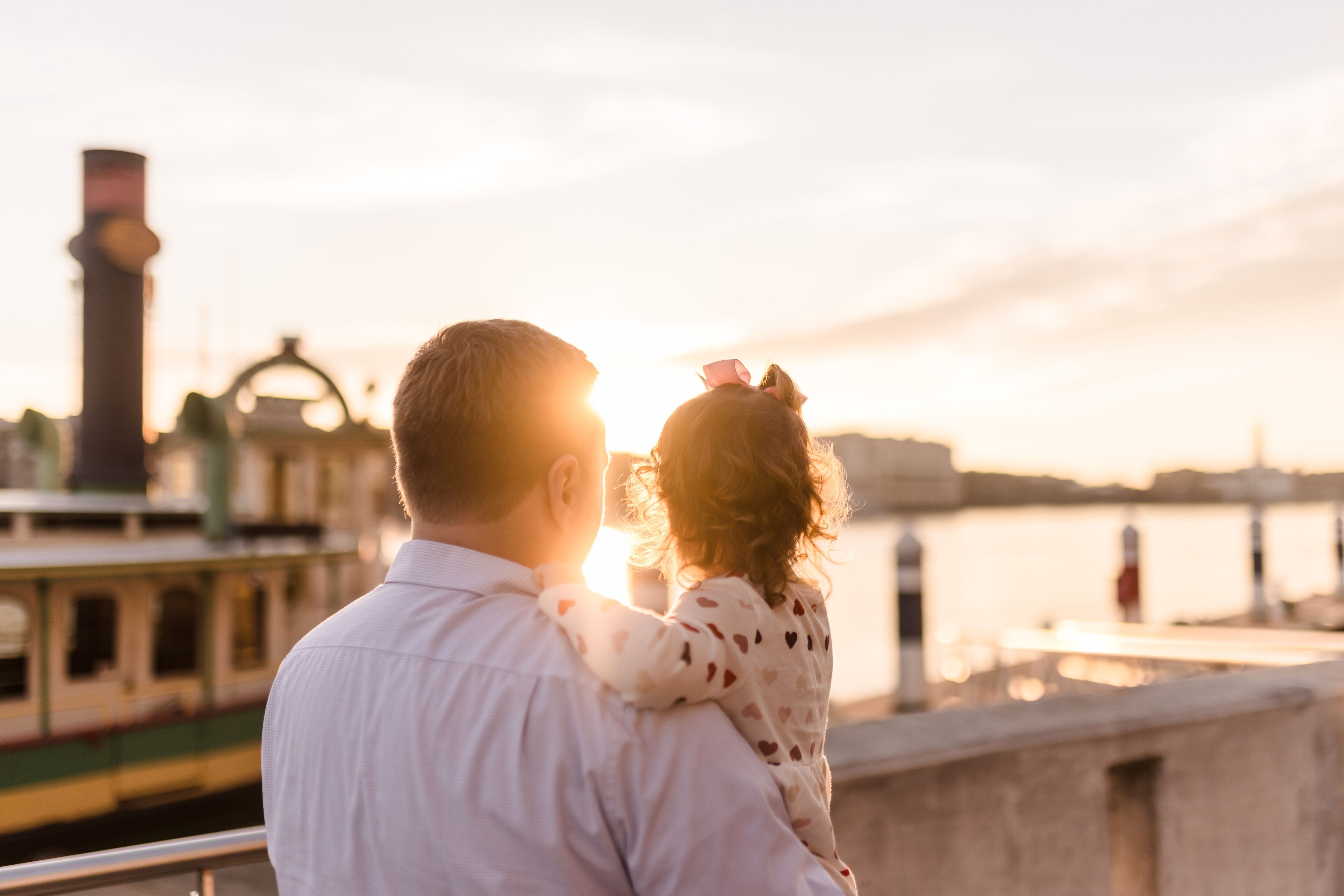 A couple embracing on a balcony overlooking a body of water with a train in the background.