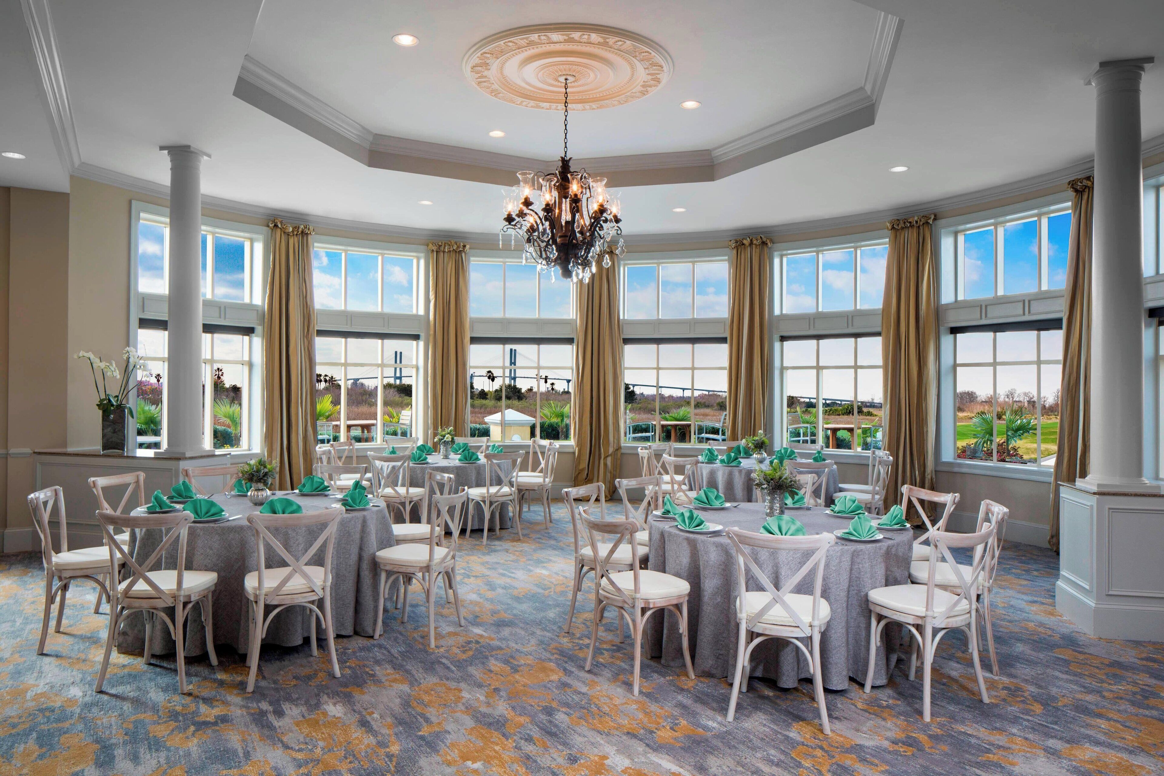A spacious room with multiple round tables and chairs, large windows with curtains, and a chandelier.