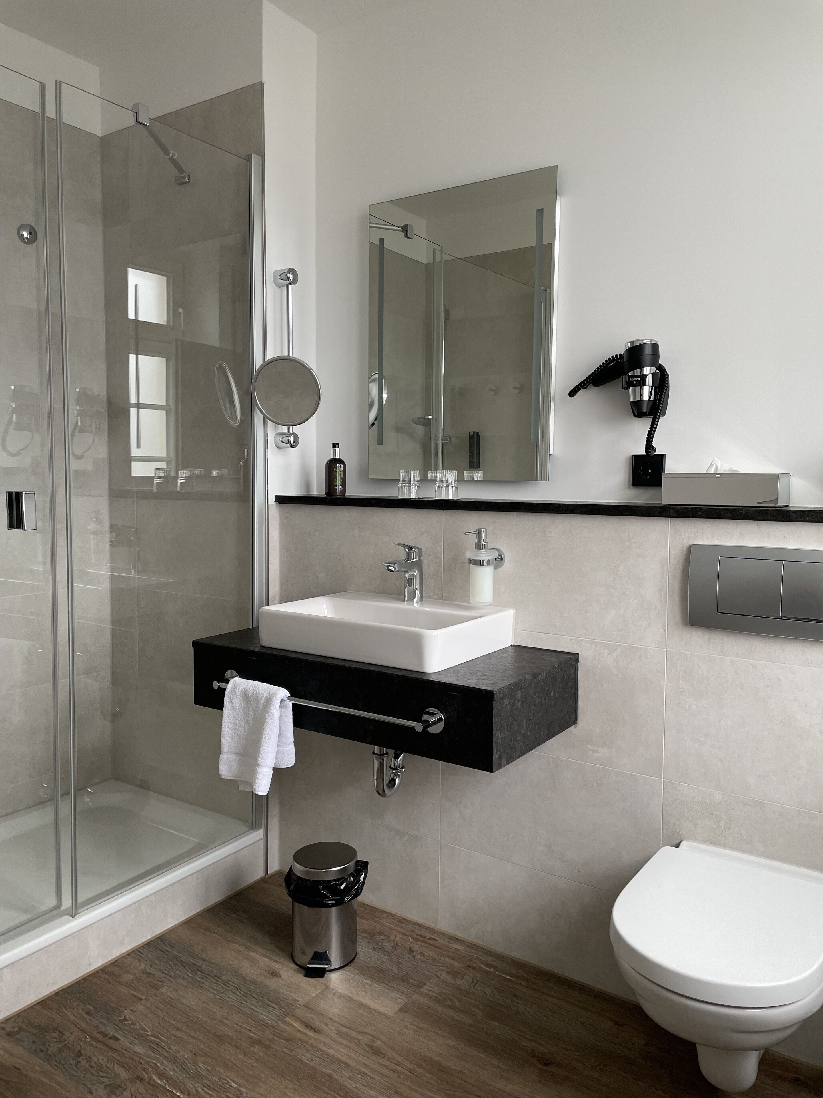 Ein modernes Badezimmer mit Duschabtrennung aus Glas, einem weißen Waschbecken mit schwarzer Arbeitsplatte, einem Spiegel, einem Fön und einer Toilette.
