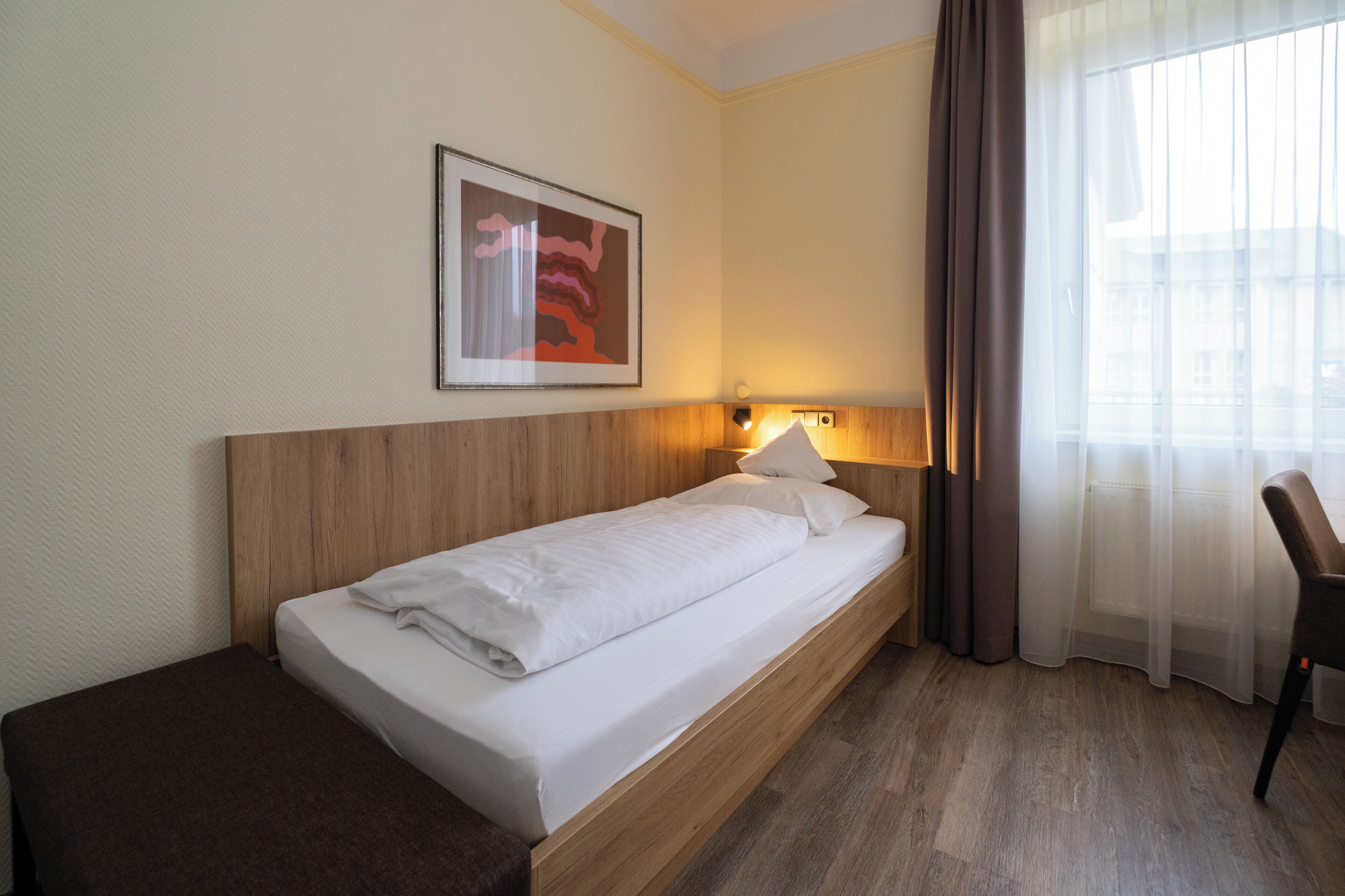 A single bed with a wooden headboard, a bedside table, a lamp, a framed picture on the wall, and a window with curtains.