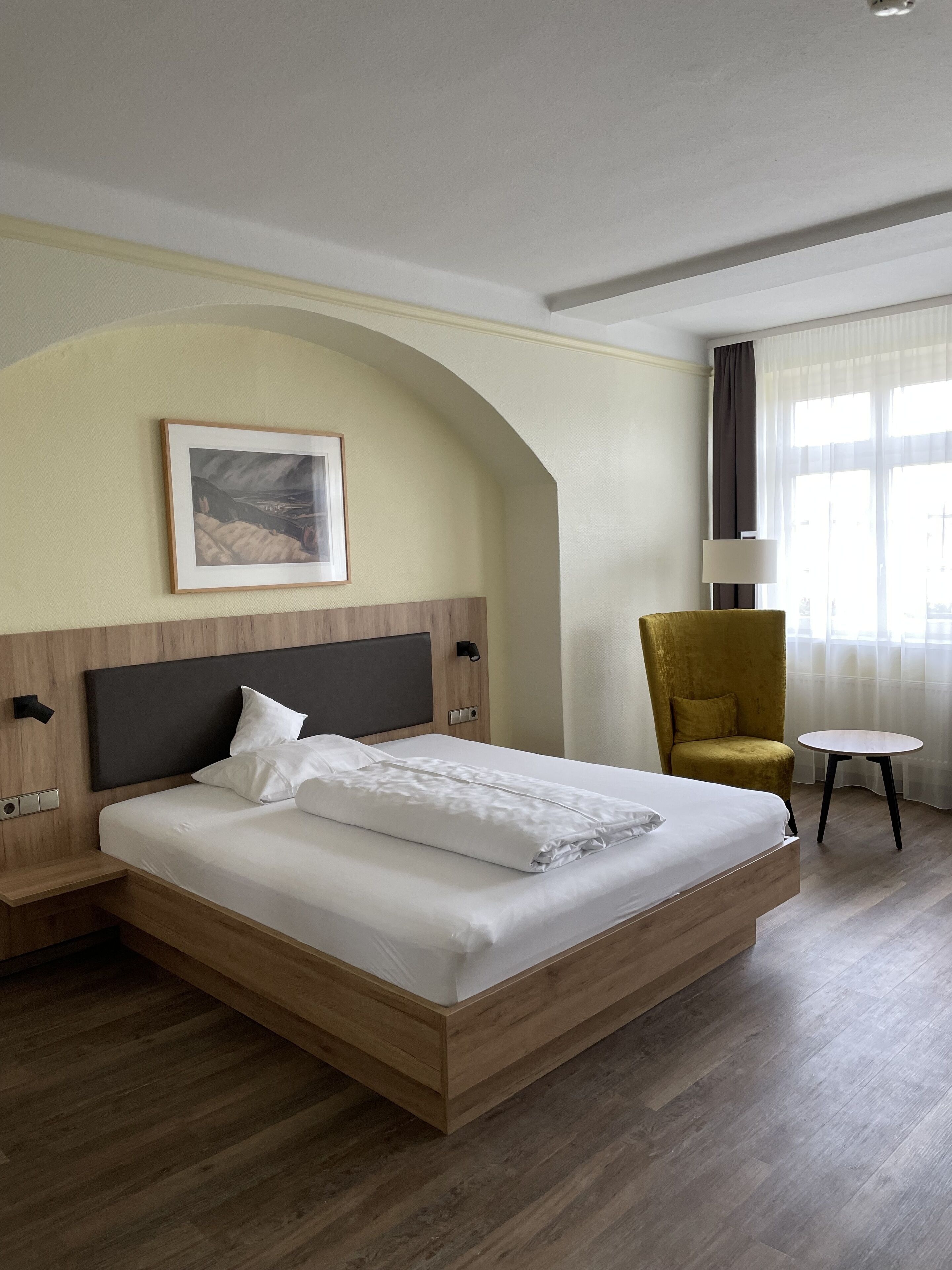 A hotel room with a large bed, a wooden headboard, a framed picture on the wall, a yellow armchair, a small round table, and a window with curtains.