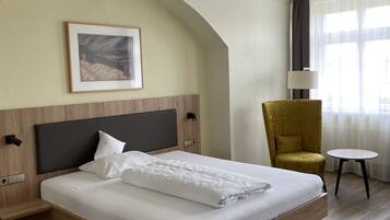 A hotel room with a large bed, a wooden headboard, a framed picture on the wall, a yellow armchair, a small round table, and a window with curtains.