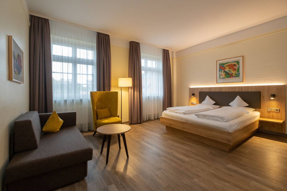 A hotel room with a bed, a sofa, a chair, a small table, and a lamp.
