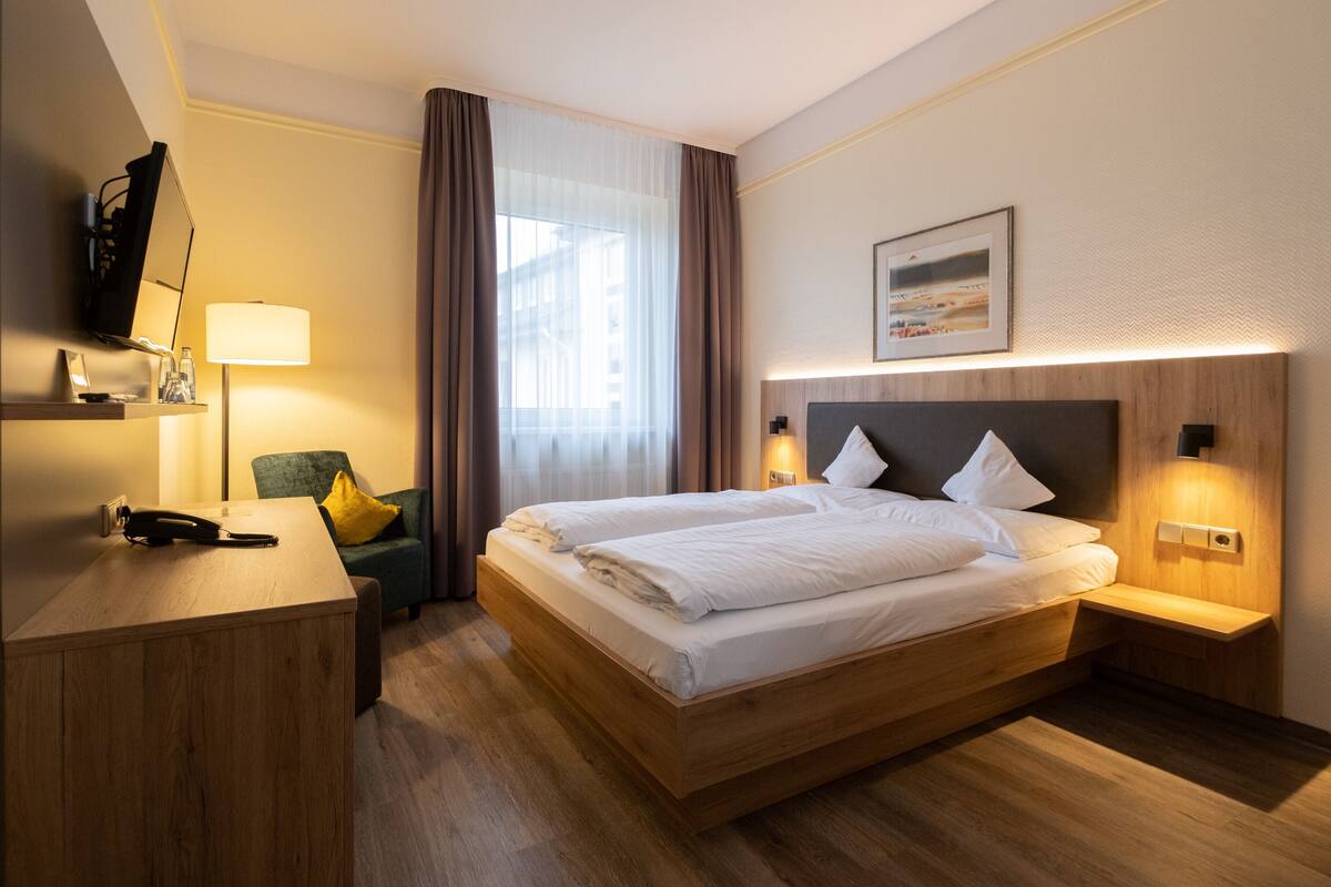 A modern hotel room with a wooden bed, a desk, a chair, a lamp, and a large window with curtains.