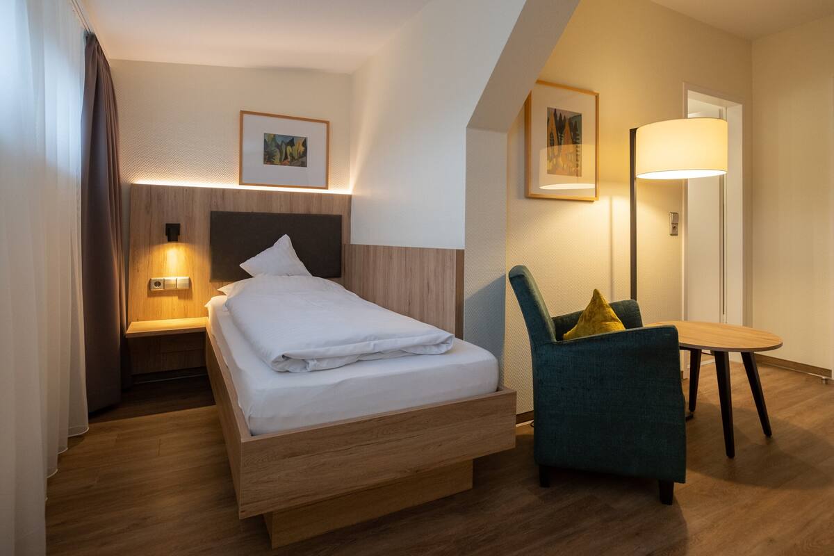 A modern hotel room with a wooden bed, a desk, a chair, and two wall-mounted lamps.