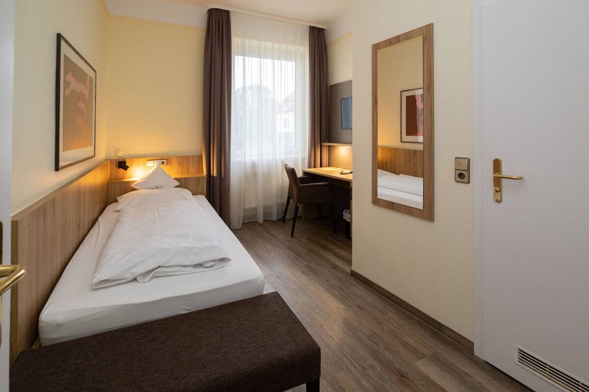 A hotel room with a bed, a desk, a chair, a bench, and a door.