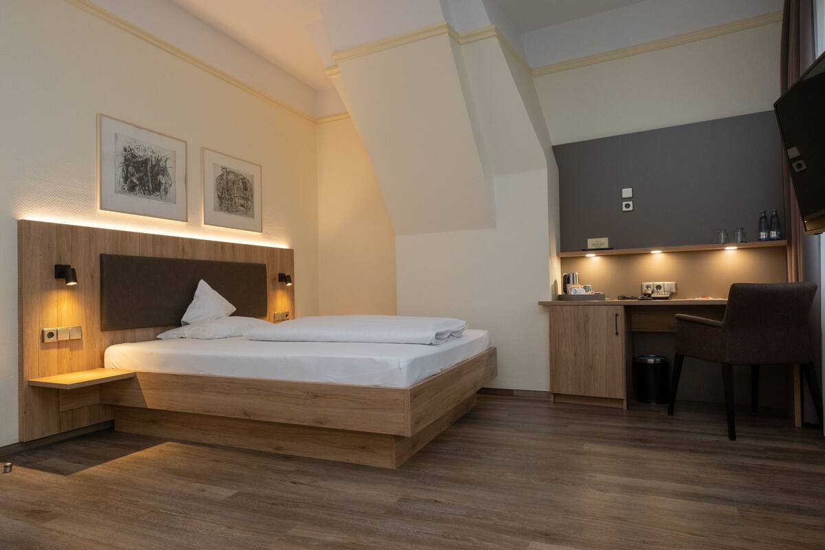 A modern hotel room with a wooden bed, a desk, and a chair. There are two framed pictures on the wall above the bed.