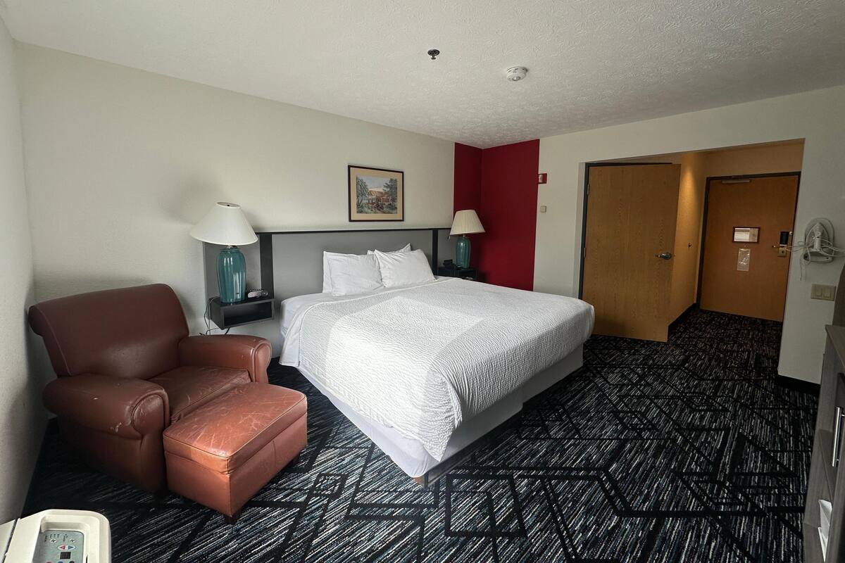 Una habitación de hotel con una cama, un sillón de cuero marrón, dos mesitas de noche con lámparas, una pared de acento roja y una puerta que da a otra habitación.