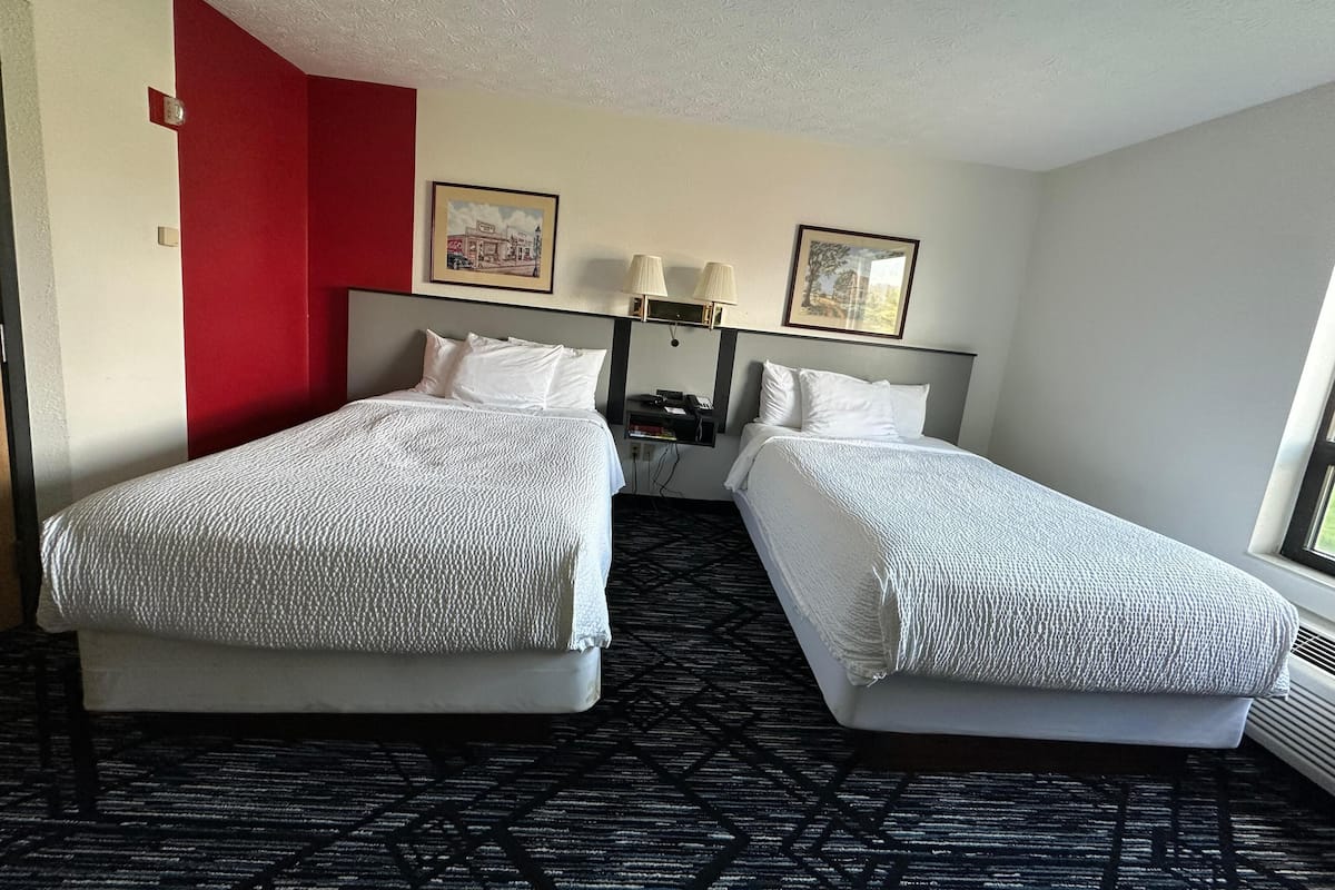 Habitación de hotel con dos camas, una pared de acento roja, un cuadro enmarcado, una lámpara y una ventana.
