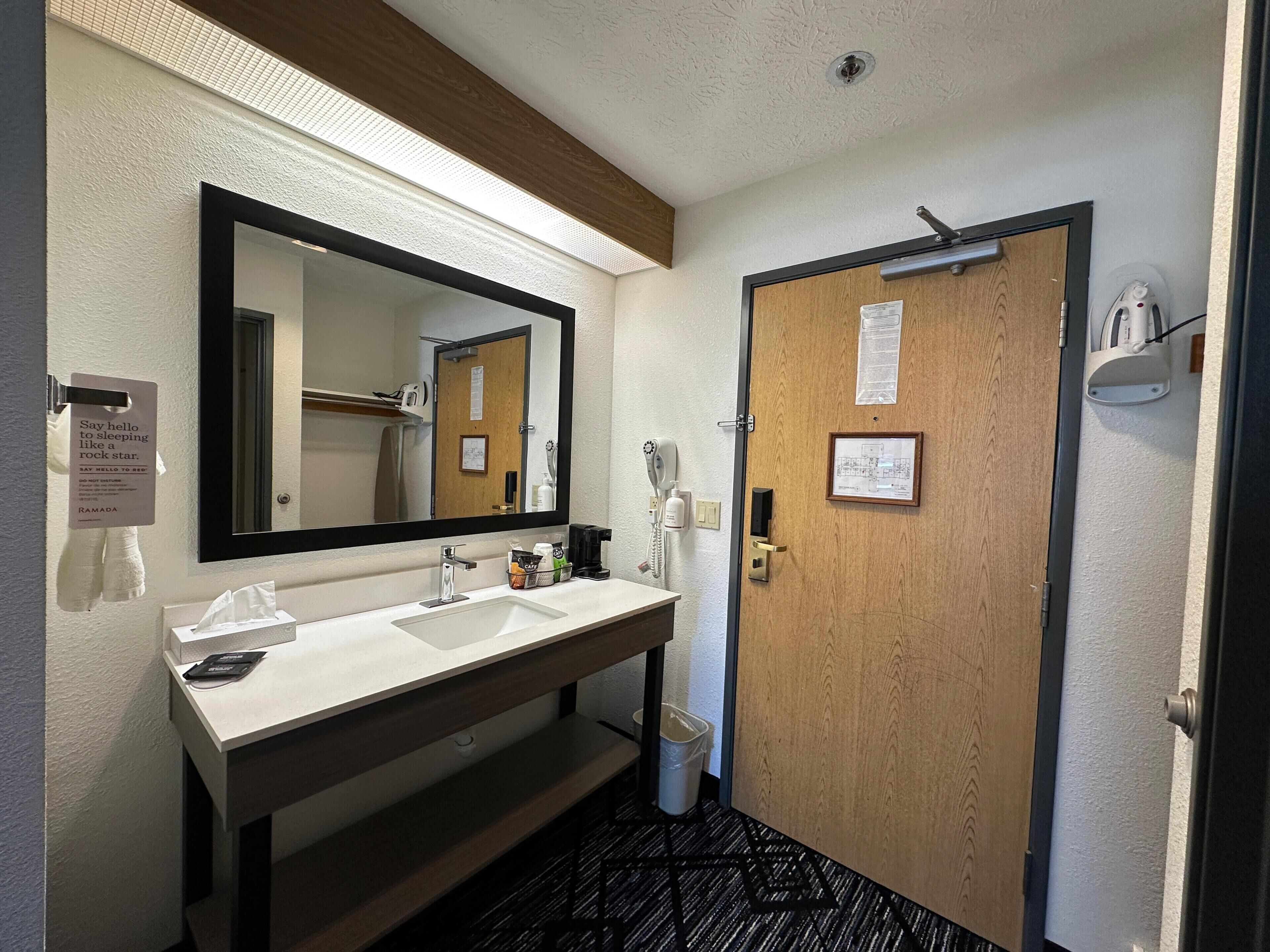 Un baño de hotel con un espejo grande, dos lavamanos, un perchero para toallas, un cubo de basura y una puerta con un letrero.