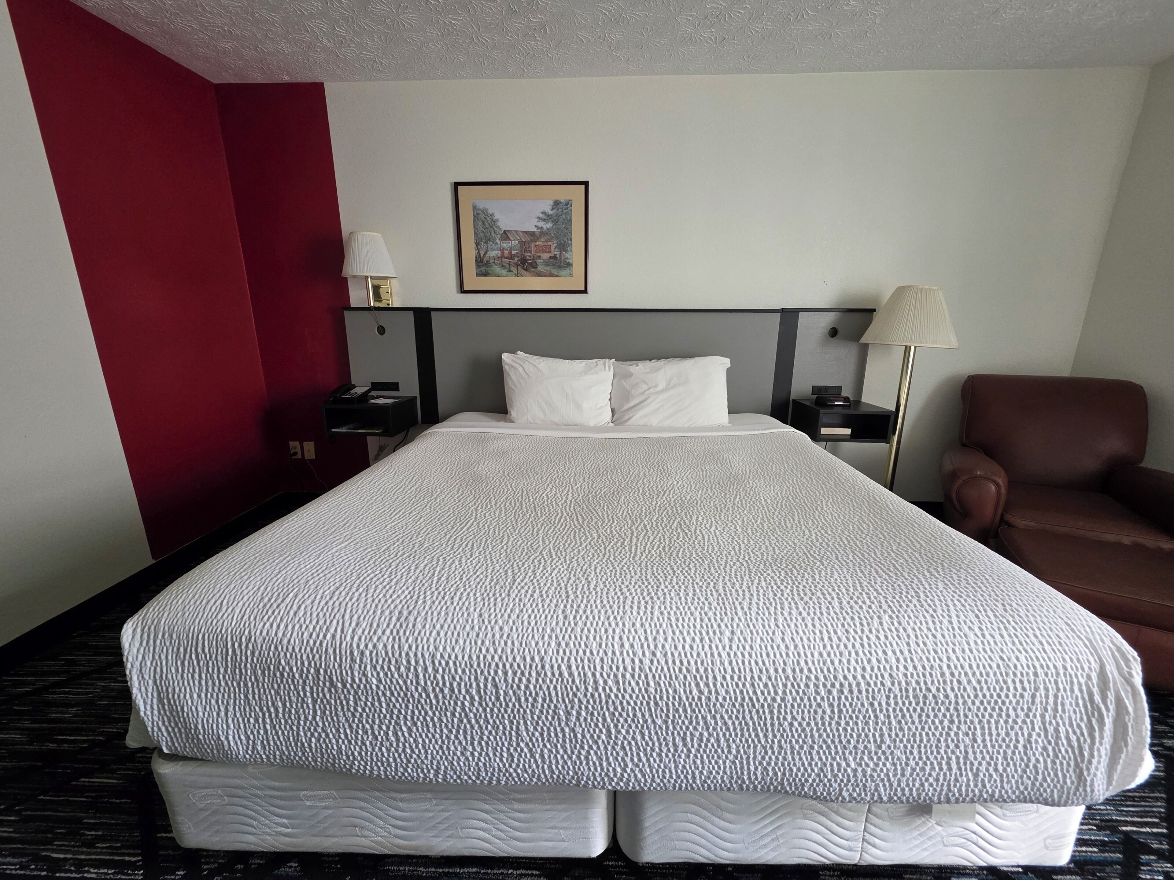 A hotel room with a large bed, a nightstand, a lamp, a chair, and a framed picture on the wall.