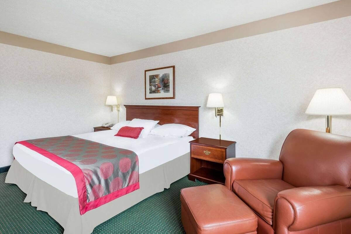 Habitación de hotel con una cama, dos mesitas de noche con lámparas, una silla y un cuadro en la pared.