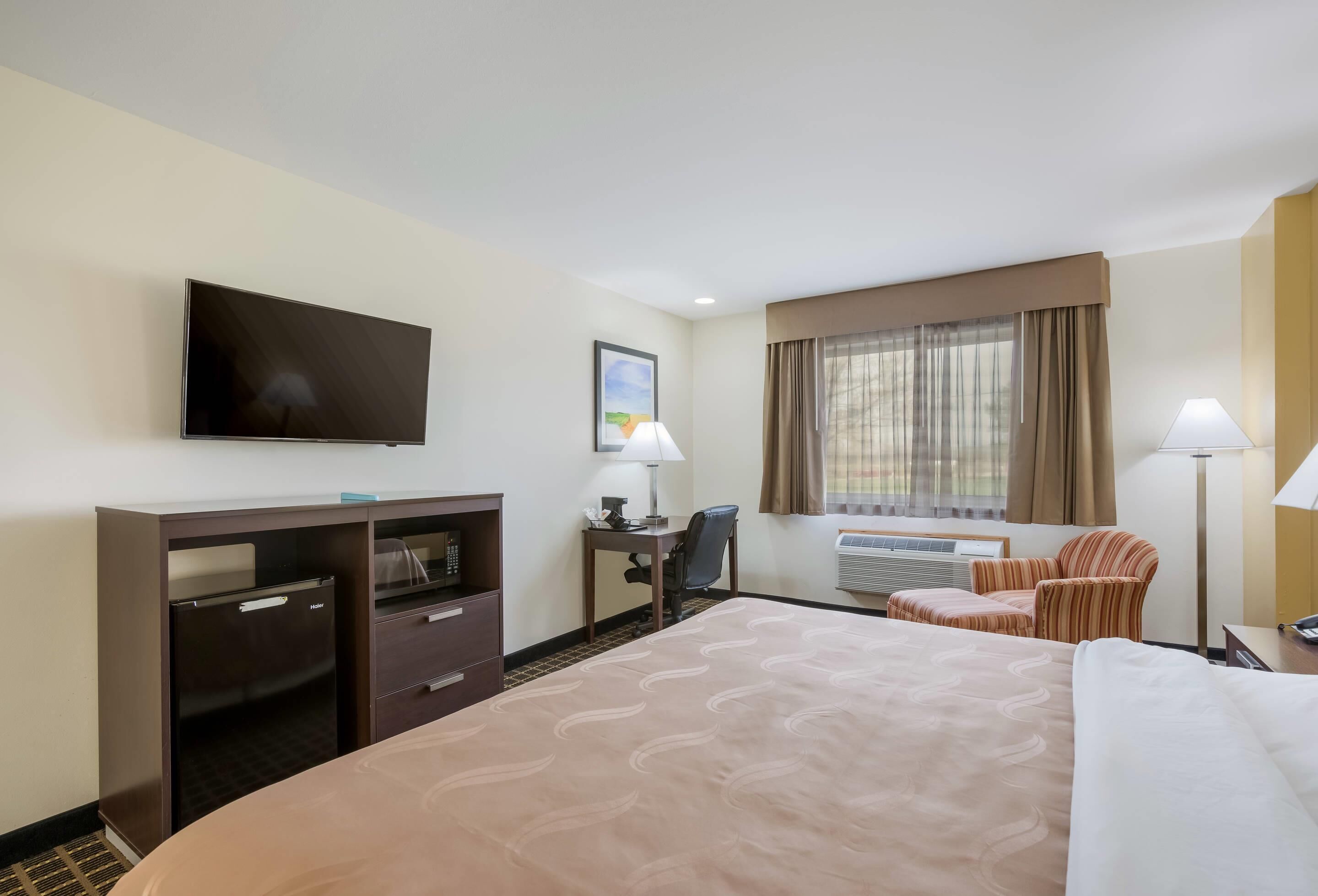 Habitación de hotel con cama, escritorio, silla, televisor, aire acondicionado y ventana con cortinas.