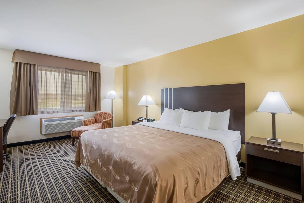 A hotel room with a bed, two bedside tables with lamps, a chair, an air conditioning unit, and a window with curtains.