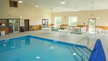 An indoor swimming pool with a blue tiled edge, surrounded by white tables and chairs, and a wooden fence.