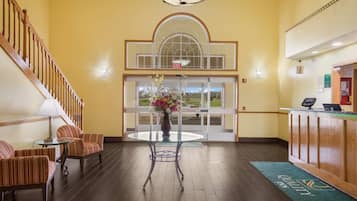 A hotel lobby with a reception desk, a staircase, a round table with chairs, and a large window with a view.