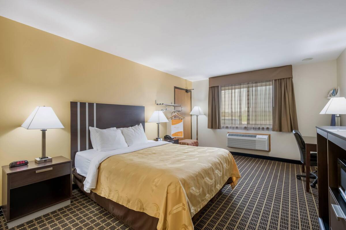 A hotel room with a bed, bedside lamps, a desk, a chair, a window with curtains, and an air conditioning unit.