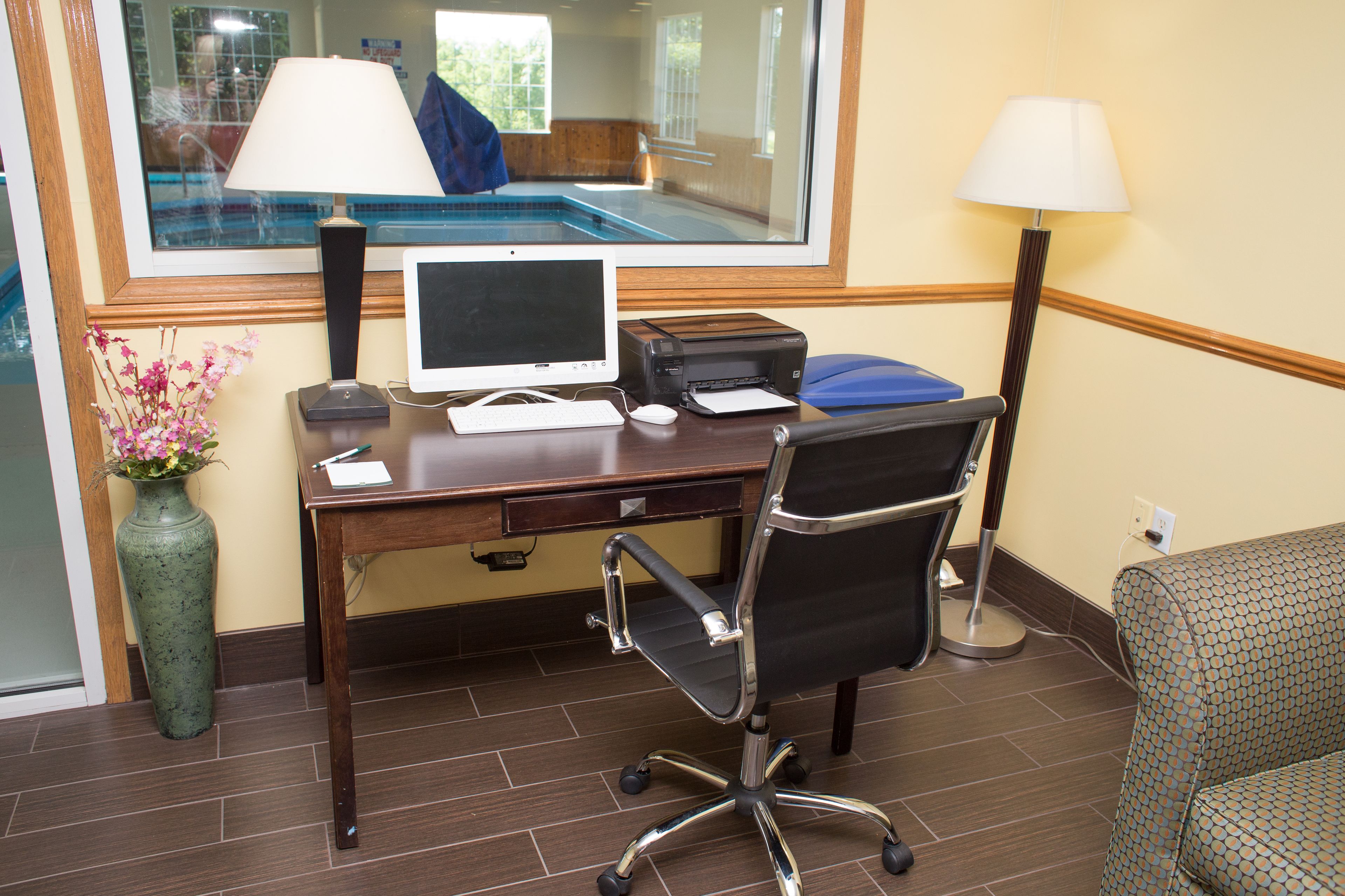 Habitación de hotel con escritorio, computadora, impresora y vista a una piscina a través de una ventana.