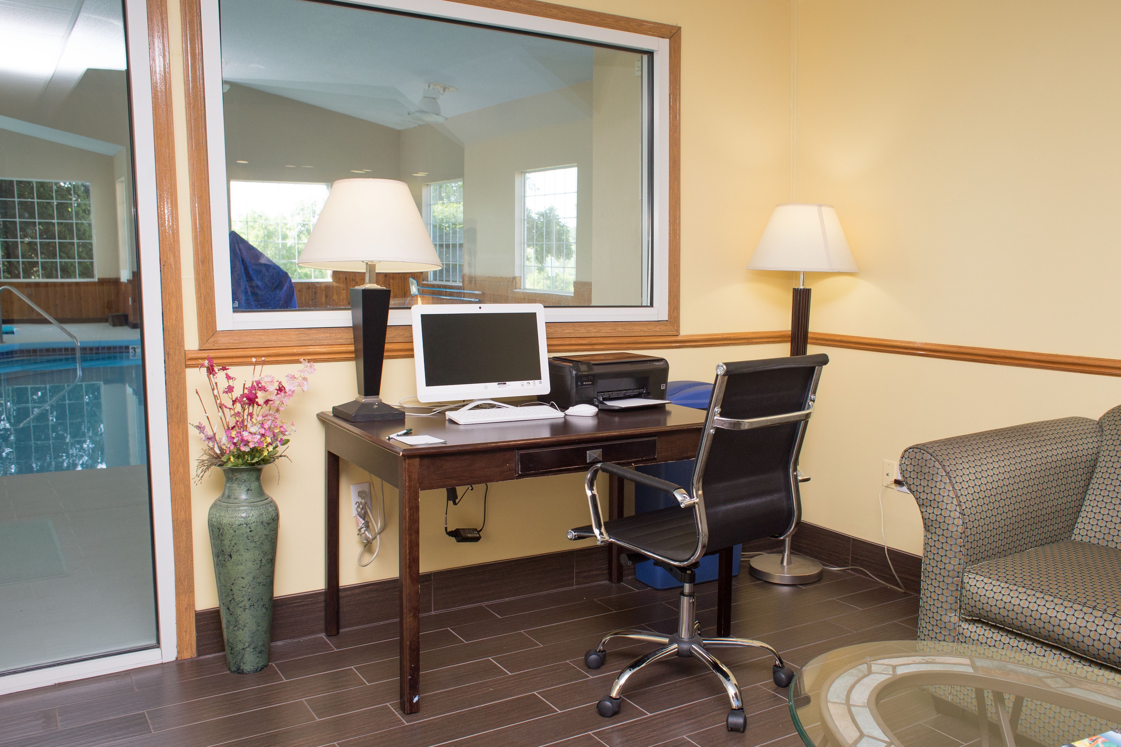 A hotel room with a desk, computer, chair, lamp, and a view of a swimming pool.
