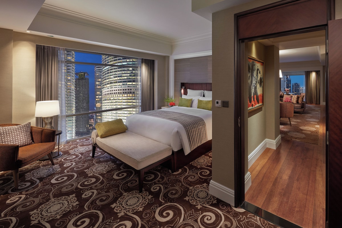 Una habitación de hotel con una cama grande, un área de descanso con una silla, vistas a un paisaje urbano a través de una ventana y una puerta que conduce a otra habitación.
