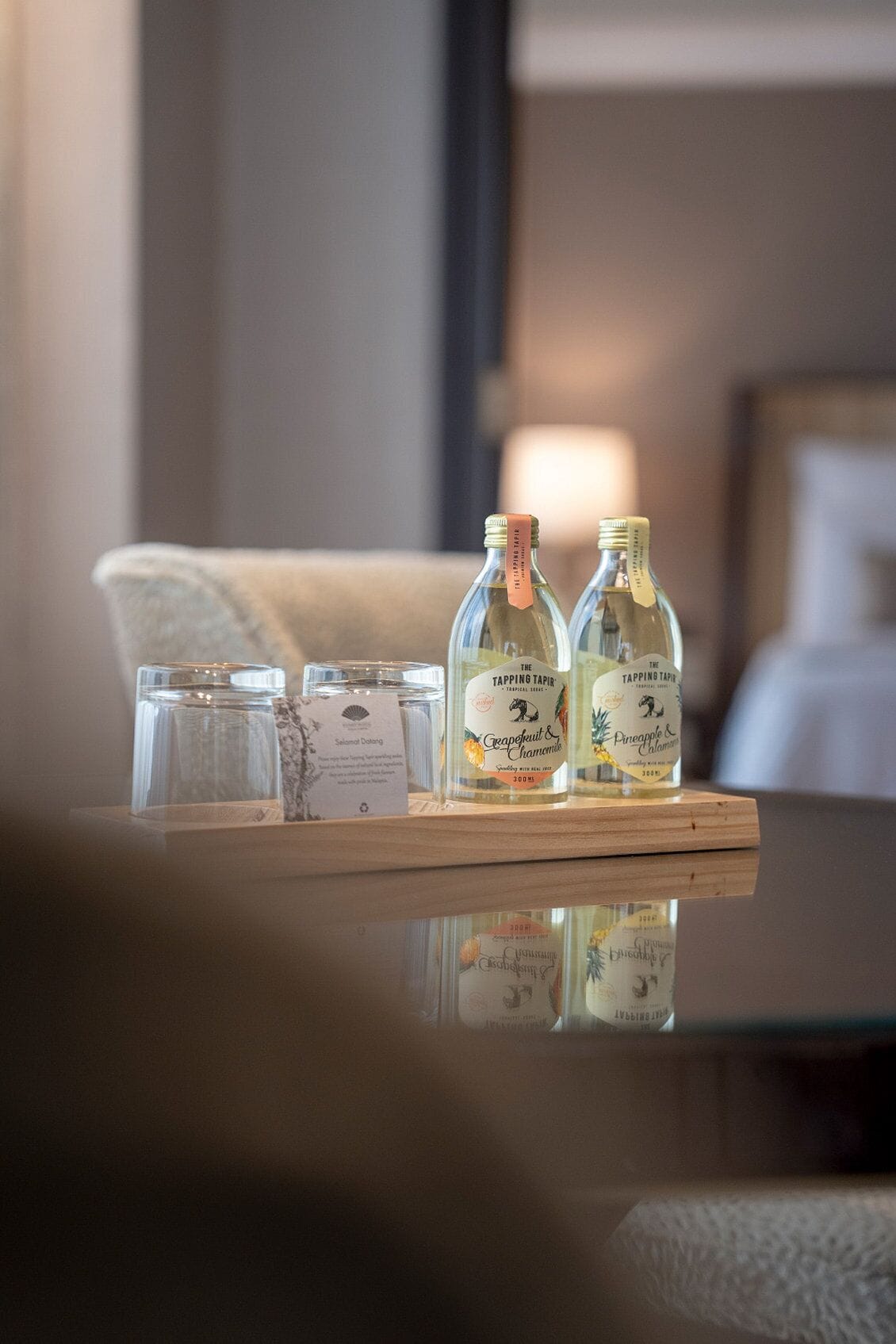 A hotel room with a wooden side table holding two bottles of 'Tapping Tapir' wine, a glass, and a small card.