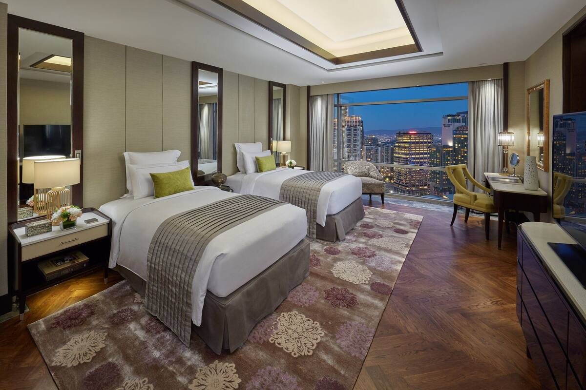 Ein Hotelzimmer mit zwei Betten, einem Schreibtisch, einem Stuhl und einem großen Fenster mit Blick auf die Stadt.