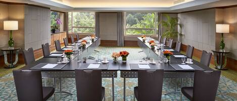 A conference room with a long table, chairs, and a view of greenery through large windows.