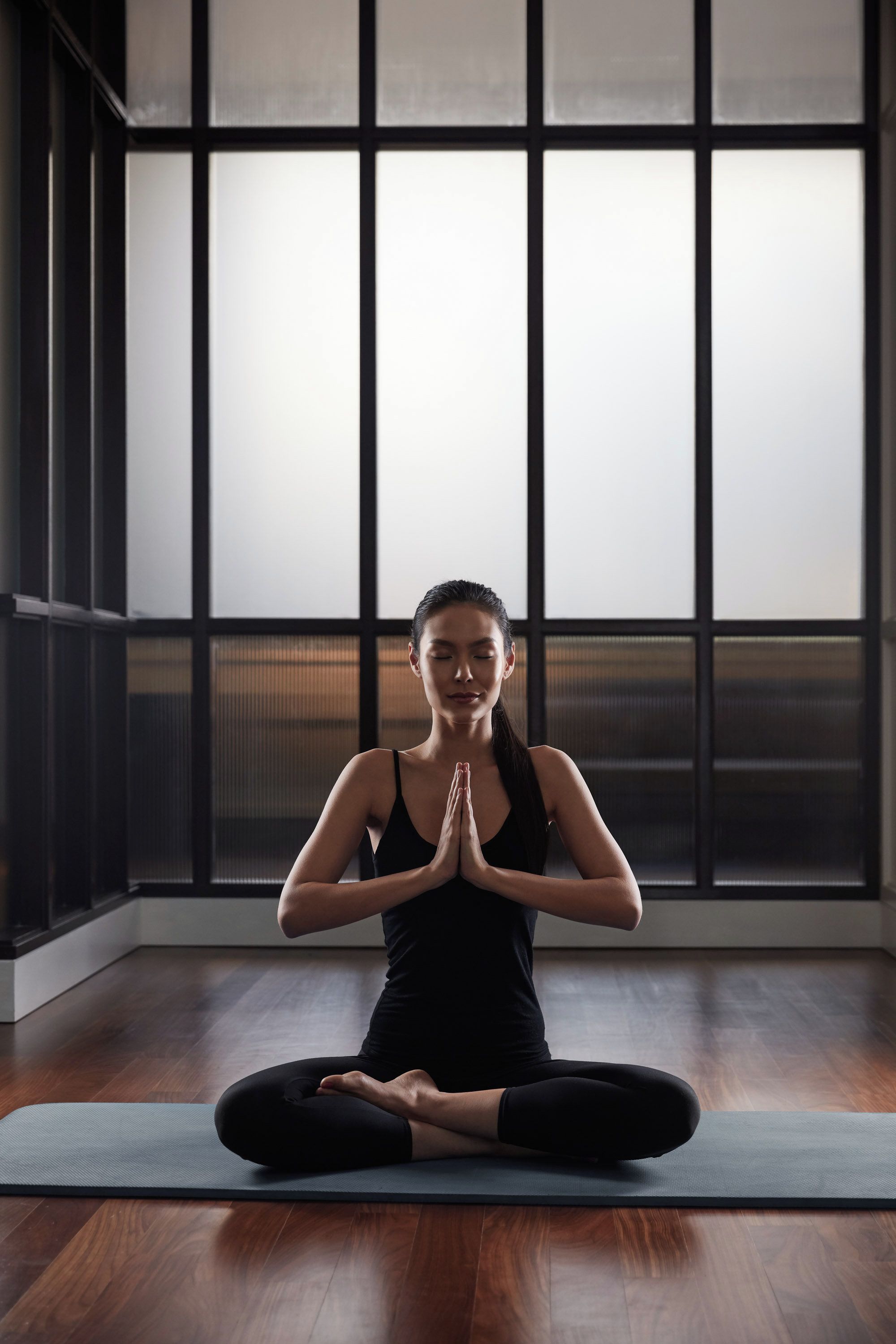 Una persona sentada en posición de meditación sobre una esterilla de yoga en una habitación con ventanas amplias y suelo de madera.