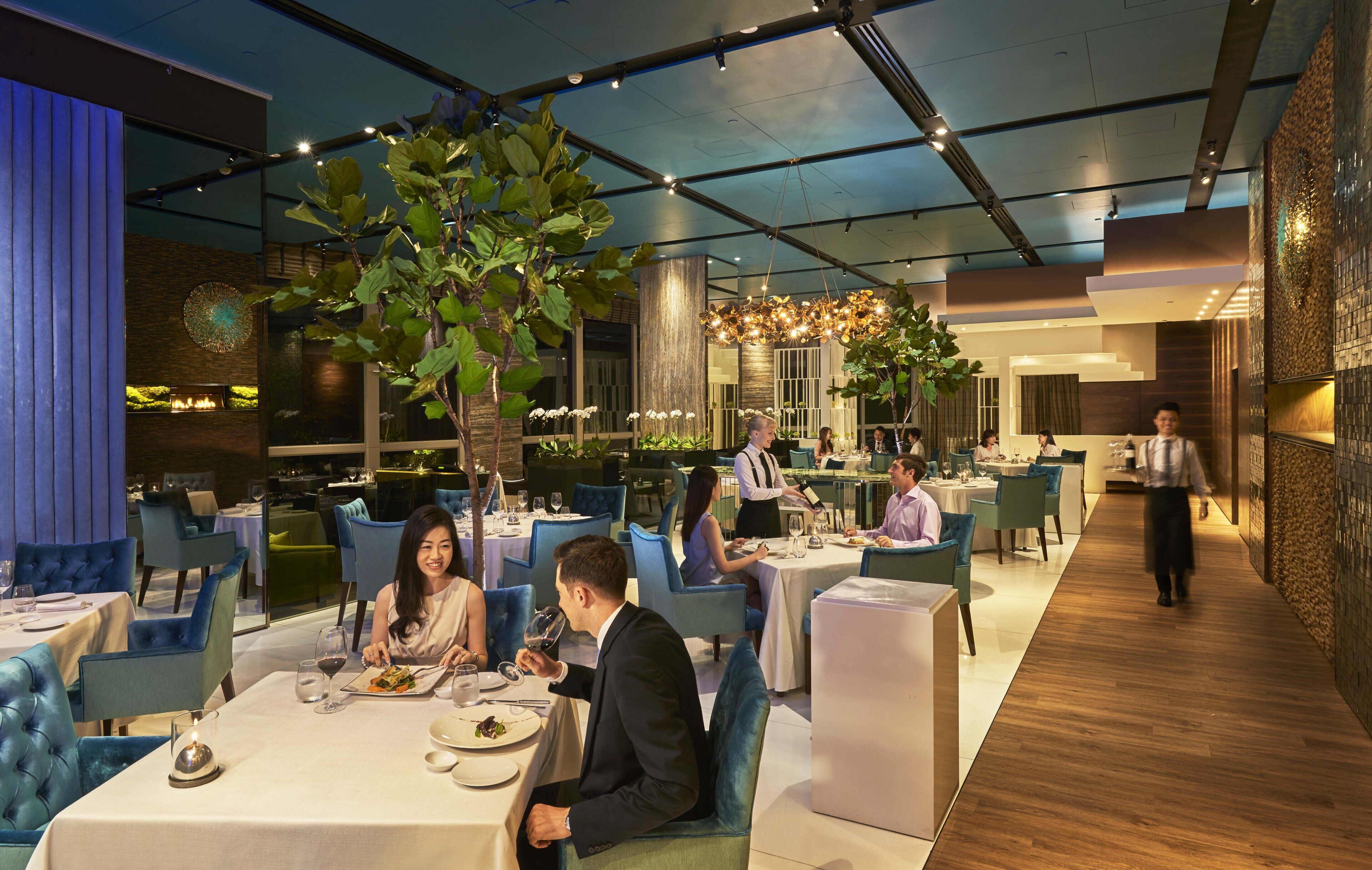 A modern restaurant with blue upholstered chairs, white tablecloths, and hanging plants.