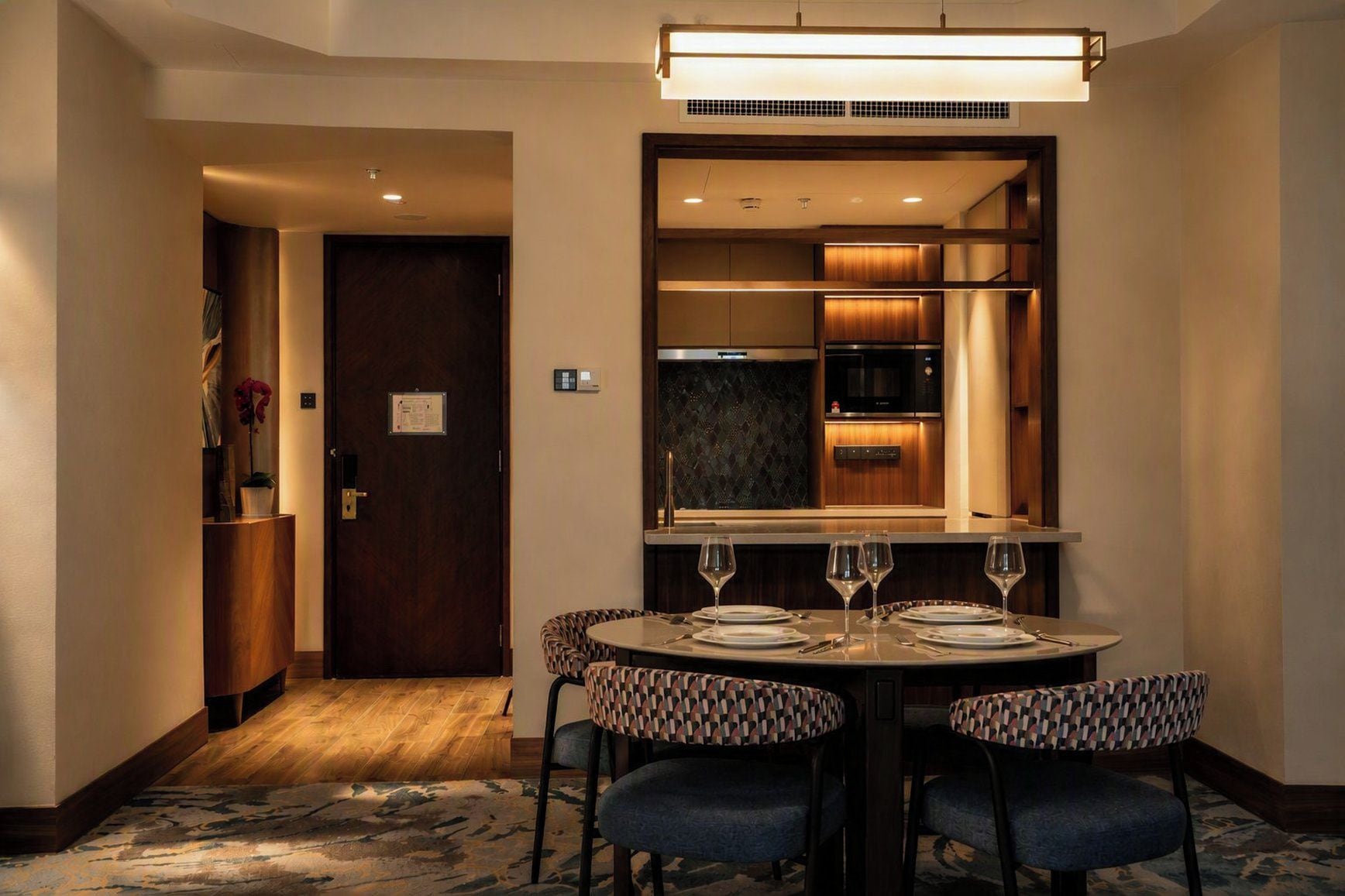 A hotel dining area with a round table set for four, a mirror above the table, and a kitchen visible through the mirror.