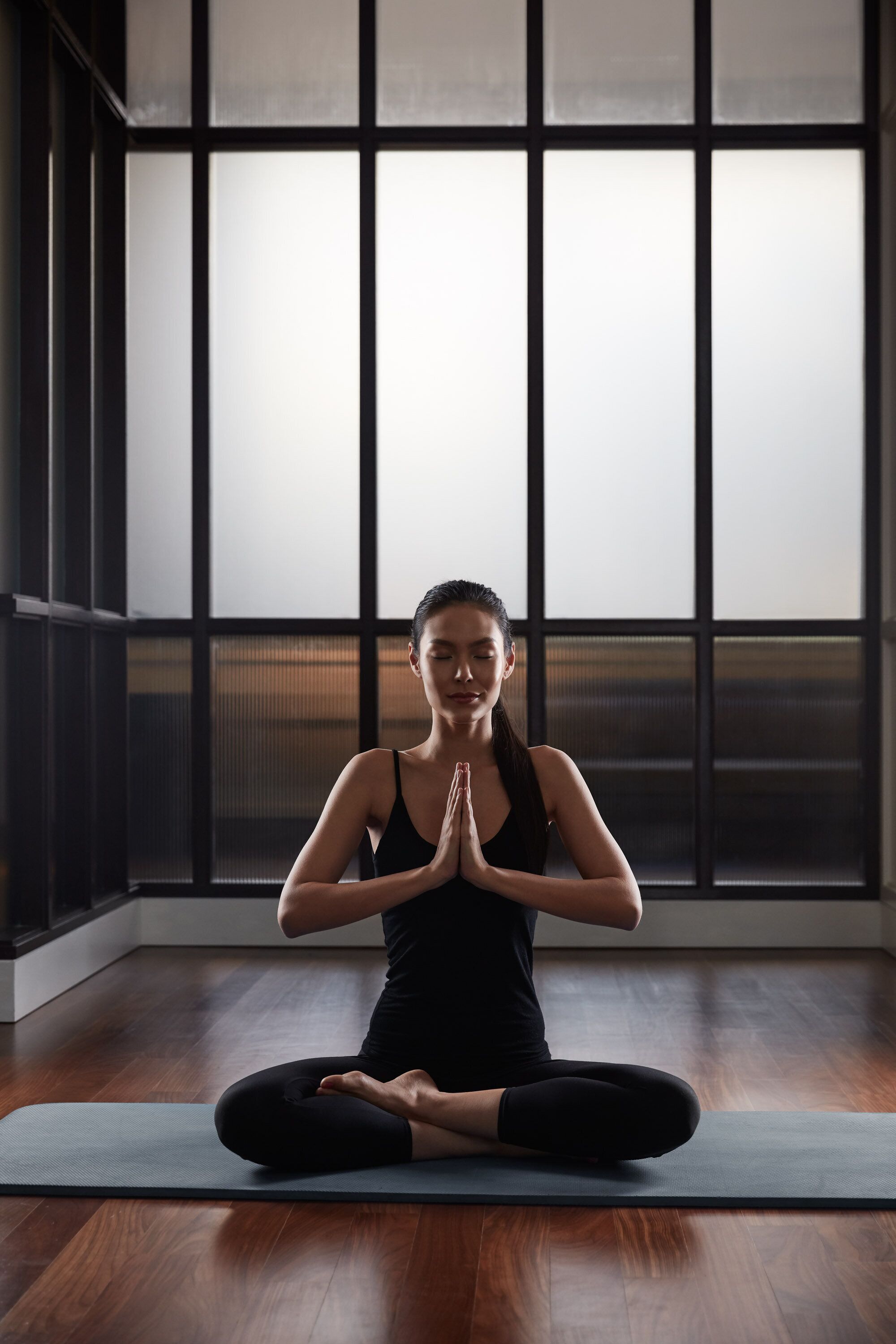 A person seated on a yoga mat in a room with large windows and wooden flooring.