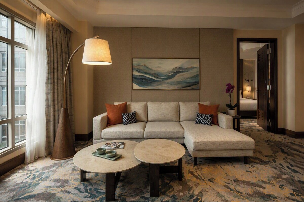 Hotelkamer met een hoekbank, twee ronde salontafels, een vloerlamp, een schilderij en uitzicht op de stad door grote ramen.