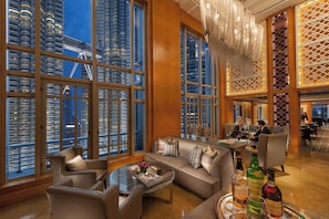 Un hall d’hôtel moderne doté d’une grande fenêtre offrant une vue sur la ville, d’un espace salon avec un canapé et des fauteuils, et d’un bar avec des bouteilles exposées.