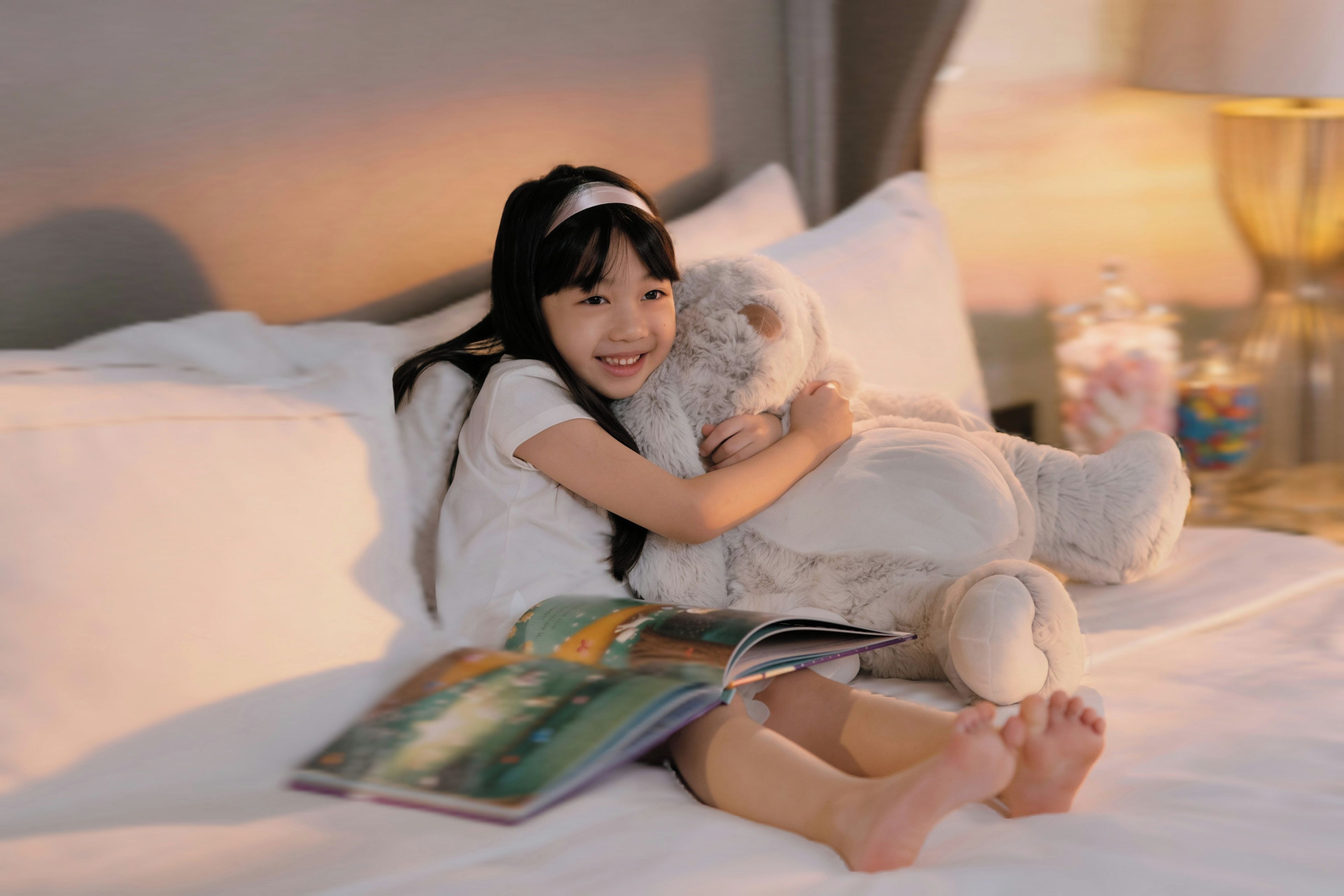 A person sitting on a bed with a book and a stuffed animal.
