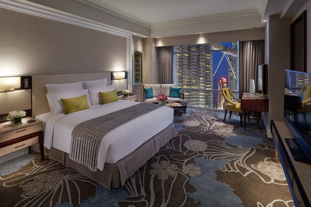 A hotel room with a large bed, a desk, a chair, and a TV. There is a view of a cityscape through the window.