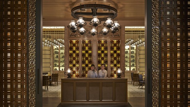 Een receptiegedeelte met een houten receptiebalie, twee personen staan erachter, en een decoratief plafond met meerdere lampen.