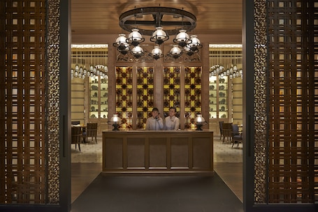 A reception area with a wooden reception desk, two people standing behind it, and a decorative ceiling with multiple light fixtures.