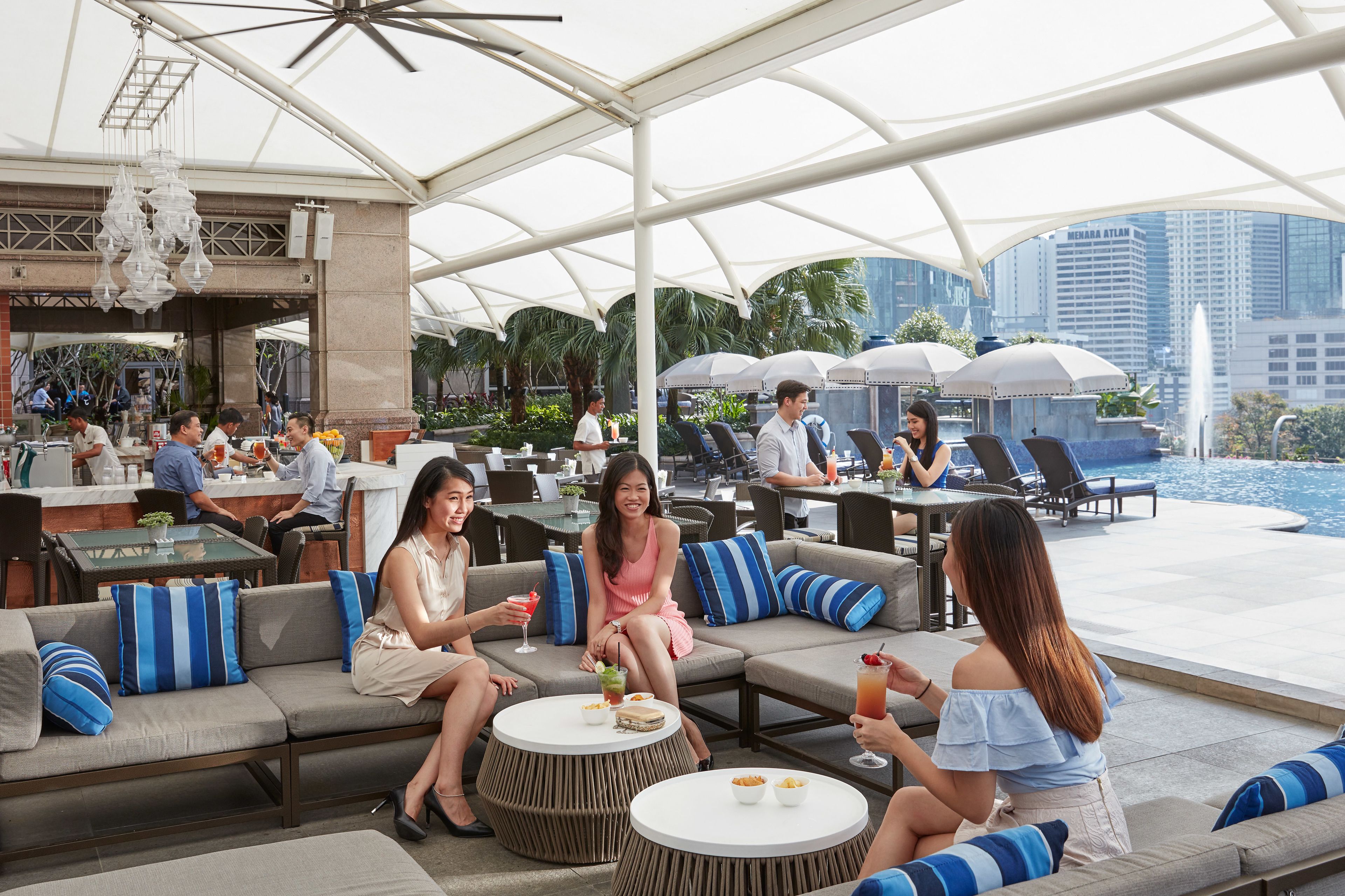 An outdoor seating area with tables and couches, a bar in the background, and a pool with umbrellas.