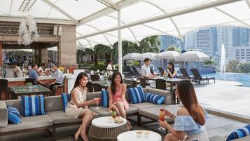 An outdoor seating area with tables and couches, a bar in the background, and a pool with umbrellas.