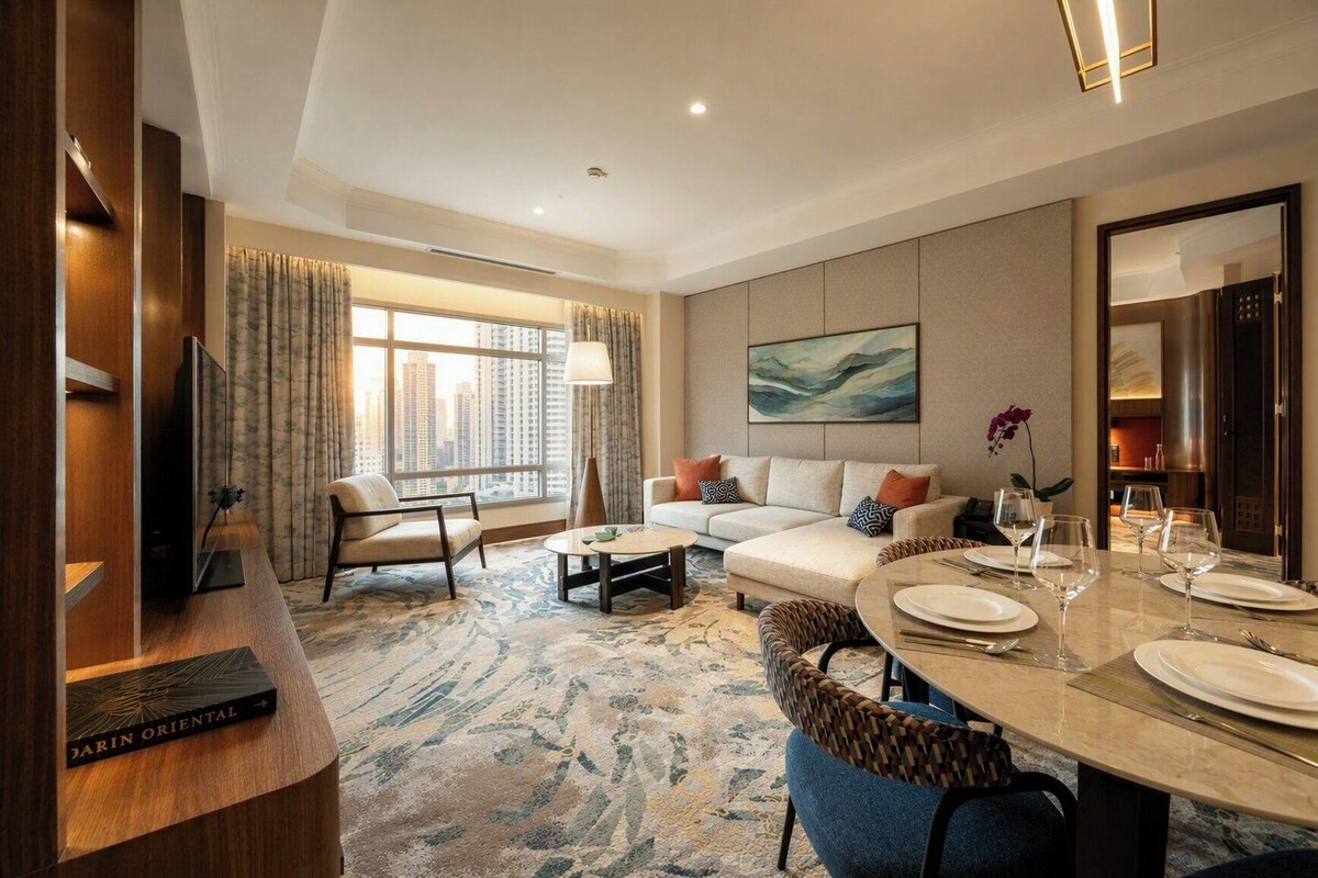Une chambre d’hôtel avec un canapé, une table à manger dressée pour quatre personnes, une télévision et une grande fenêtre avec des rideaux.