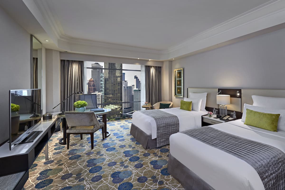 Ein Hotelzimmer mit zwei Betten, einem Schreibtisch mit Computer, einem Stuhl und Blick auf die Stadt.