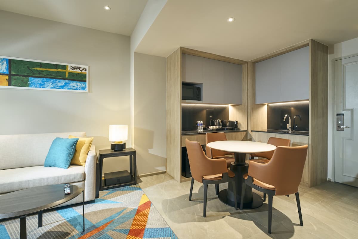 Een moderne hotelkamer met een bank, een eettafel met stoelen, een keukenhoek met een magnetron en waterkoker, en een schilderij aan de muur.